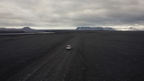 Toma-De-Un-Dron-De-Un-Automóvil-Plateado-Que-Conduce-Por-Una-Carretera-En-Un-Campo-De-Lava-En-Islandia-Con-La-Cámara-Dando-Vueltas-Y-Montañas-En-El-Fondo-4k
