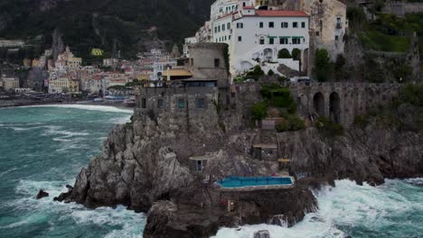 Luxury-pool-in-cliff-at-Amalfi-coast,-aerial-view-of-waves-banging-on-shore