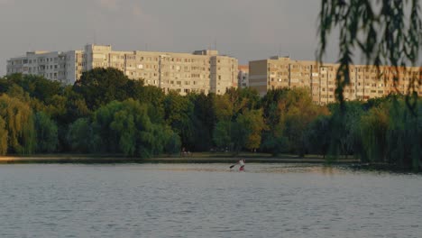 Ein-Kajak,-Das-Bei-Sonnenuntergang-Auf-Einem-See-Mit-Einigen-Alten-Kommunistischen-Gebäuden-Und-Bäumen-Im-Hintergrund-Vorbeifährt