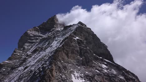 Ascending-aerial-view-from-drone-of-The-Matterhorn,-famous-peak-top-of-Mount-Cervino-with-bright-blue-sky-on-one-side-and-soft-white-clouds-on-other