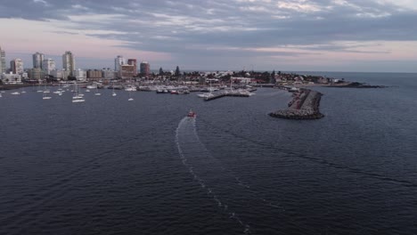 Cinematic-aerial-circling-shot-of-red-fishing-motorboat-entering-the-city-harbor-of-Punta-del-Este-in-Uruguay-with-skyline-and-skyscrapers-in-background