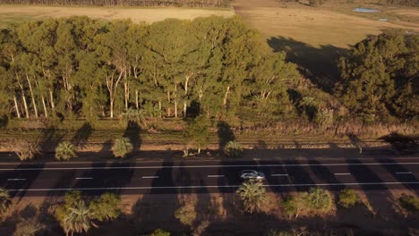 Grey-car-driving-along-rural-route-at-sunset-in-Uruguay,-aerial-drone-pov