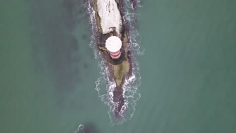 Aerial-drone-flight-giving-a-birdseye-view-of-the-red-and-white-lighthouse-at-The-Needles-in-the-Isle-of-Wight-showing-crashing-waves-hitting-the-rocks-below