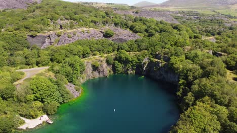 Aerial-view-Dorothea-slate-mining-quarry-woodland-in-Snowdonia-mountain-valley-with-gorgeous-shimmering-blue-lake