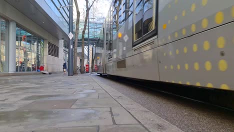 A-Yellow-Metrolink-tram-travelling-down-a-street-and-under-a-glass-footbridge-with-people-passing-by-in-Manchester-City-Centre