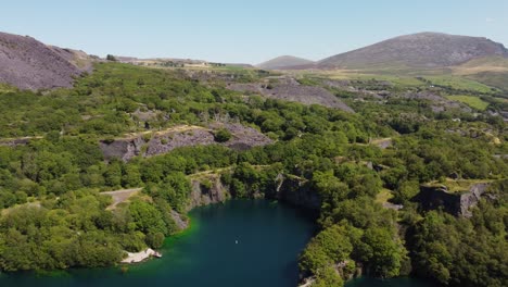 Aerial-view-Dorothea-slate-mine-quarry-ruins-woodland-in-Snowdonia-valley-with-gorgeous-shimmering-blue-lake