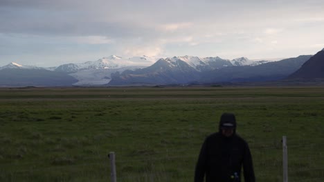 Hombre-Caminando-A-La-Cámara-Con-Praderas-De-Islandia-Y-Montañas-En-El-Fondo