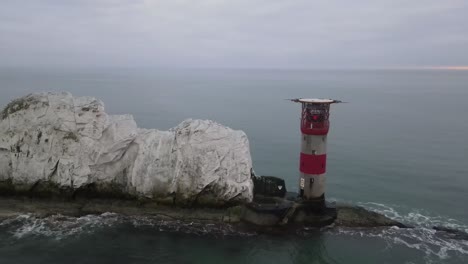 Aerial-drone-flight-around-the-red-and-white-Lighthouse-at-The-Needles-in-the-Isle-of-Wight-showing-a-detailed-view-of-the-white-cliffs-and-waves-crashing-against-the-rocks