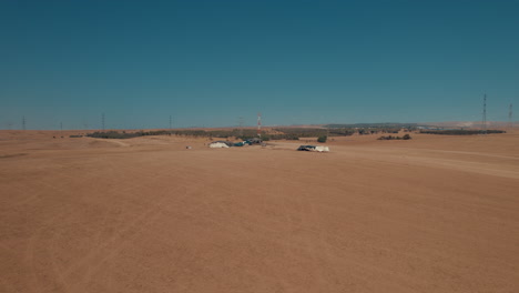 Bedouin-tents-in-an-arid-and-remote-area,-on-a-dry-sand-field-off-the-grid,-near-large-power-lines-and-an-animal-feed-factory-parallax-shot