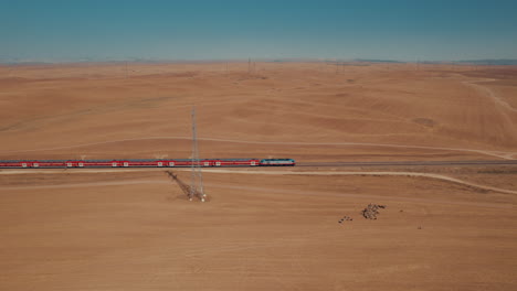 Aerial-shot-of-a-red-passenger-train-in-a-remote-and-desert-area,-near-large-electricity-poles,-dry-land-without-crops,-on-the-ground-there-is-a-female-shepherd-with-sheeps