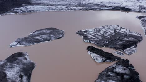 Icebergs-with-black-ash-floating-in-glacial-lagoon-at-Kvíarjökull-glacier,-aerial