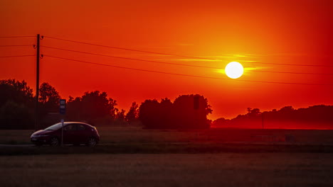 A-car-parked-along-a-countryside-road-as-a-fiery-sun-descends-behind-a-copes-of-trees---time-lapse