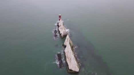 Aerial-drone-flight-in-the-Isle-of-Wight-looking-down-on-the-red-and-white-Lighthouse-at-The-Needles-with-waves-crashing-against-the-white-cliffs