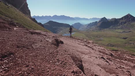 Back-view-of-young-hiker-walking-on-red-landscape-towards-a-lake-and-enjoy-the-views-of-Pyrenees-mountains