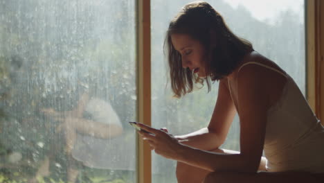 A-young-half-naked-woman-sitting-on-a-bed-in-the-morning-sun,-texting-on-her-cell-phone