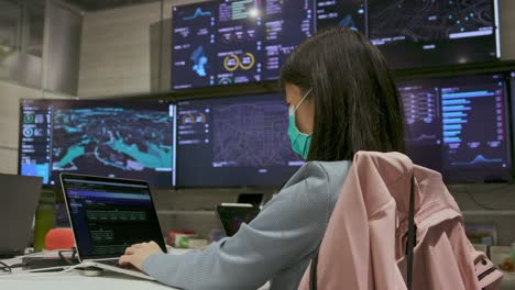Asian-woman-in-an-office-wearing-a-mask-while-working-on-a-computer-with-big-screens-in-the-background