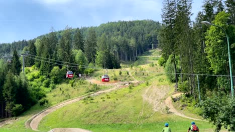 Red-gondolas-passing-each-other-on-cableway,-lush-forest-on-mountainside