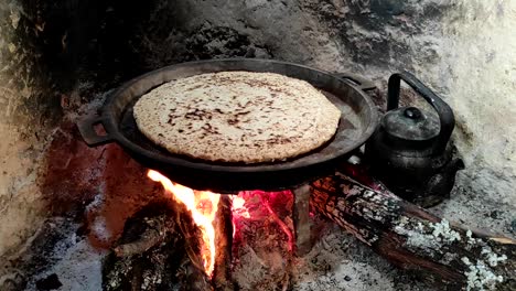 Close-up-of-Cooking-bread-in-a-cast-iron-skillet-resting-on-trivets,-over-an-oak-wood-fire-in-a-large-stone-fireplace-in-a-traditional-kitchen-4