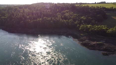Aerial-view-above-foaming-sparkling-sunset-river-flowing-alongside-Menai-Straits-woodland-countryside
