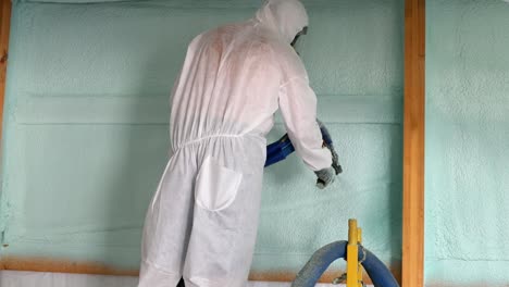Worker-in-respirator-and-Tyvek-suit-sprays-second-closed-cell-foam-insulation-on-the-house-wrap-and-studs-of-newly-constructed-exterior-wall