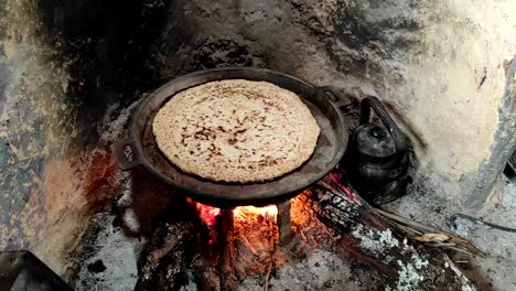 Close-up-of-Cooking-bread-in-a-cast-iron-skillet-resting-on-trivets,-over-an-oak-wood-fire-in-a-large-stone-fireplace-in-a-traditional-kitchen