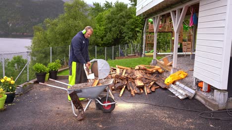 Male-european-man-using-electrical-saw-to-cut-wood-and-prepare-for-winter-outside-his-house---Wooden-pine-planks-cut-and-falling-into-wheelbarrow---Sea-and-trees-in-background