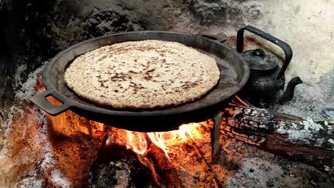 Close-up-of-Cooking-bread-in-a-cast-iron-skillet-resting-on-trivets,-over-an-oak-wood-fire-in-a-large-stone-fireplace-in-a-traditional-kitchen-1