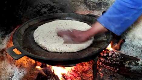 Close-up-of-Cooking-bread-in-a-cast-iron-skillet-resting-on-trivets-and-woman's-hand-rolling-out-pastry-into-a-circle,-over-a-wood-fire-in-a-fireplace
