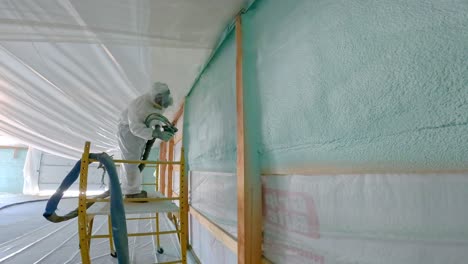 Man-in-respirator-and-Tyvek-suit-sprays-closed-cell-foam-insulation-on-exterior-wall-for-new-construction-while-standing-on-scaffolding