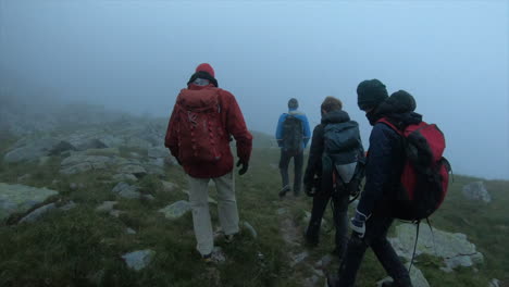 A-group-of-mountain-climbers-walk-in-a-morning-fog-with-backpack-on-grass-and-rocks,-swiss-alps-adventure