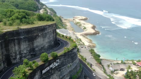 Ein-Auto-Fährt-über-Die-S-förmige-Straße,-Die-Den-Hügel-Durchschneidet,-Und-Der-Melastische-Strand-Von-Uluwatu-Bali-Ist-Von-Der-Hügelspitze-Aus-Zu-Sehen