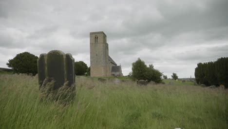 Wind-blowing-through-an-overgrown-graveyard-showing-an-ancient-medieval-church-in-the-background-and-a-dramatic-overcast-sky
