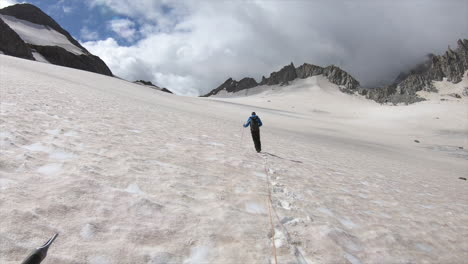 Un-Montañero-Camina-Sobre-Un-Glaciar-Cubierto-De-Nieve-Junto-A-Cumbres-Rocosas-Y-Afiladas,-Se-Le-Ata-Una-Cuerda-Para-Mantener-A-Todos-Juntos,-Expedición