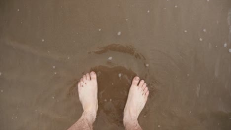 A-mans-feet-stood-on-the-beach-as-the-tide-comes-in-and-water-laps-at-the-shore