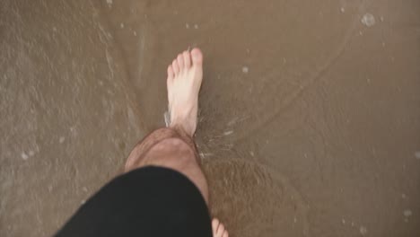 A-caucasian-mans-feet-walking-on-a-beach-as-the-tide-periodically-comes-in
