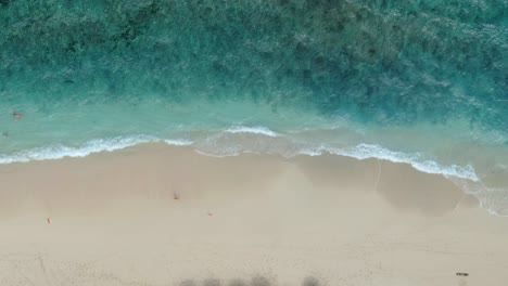 Drone-footage-shows-a-beach-being-pounded-by-many-little-waves-that-turn-white-and-foamy,-and-some-people-swimming-in-the-clear-blue-water