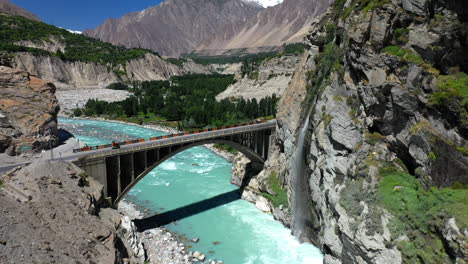 Drone-shot-of-turquoise-blue-water-river-flowing-under-a-bridge-in-Karakoram-mountain-range-along-Karakoram-highway,-moving-towards-bridge-with-traffic
