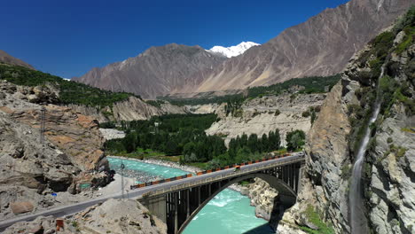 Drone-shot-of-turquoise-blue-water-river-flowing-under-a-bridge-in-Karakoram-mountain-range-along-Karakoram-highway,-slowly-revealing