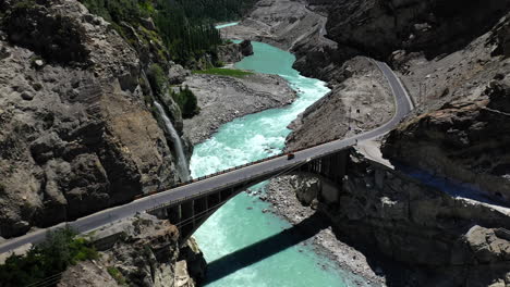 Drone-shot-of-turquoise-blue-water-river-flowing-under-a-bridge-in-Karakoram-mountain-range-along-Karakoram-highway,-revealing