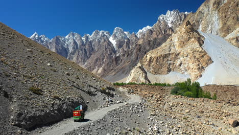 Cinematic-drone-shot-of-a-tuk-tuk-on-a-gravel-path-on-the-Karakoram-Highway-Pakistan-with-the-passu-cones-in-the-distance,-descending-aerial-shot