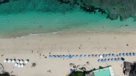 Luftbild-Des-Melasti-Strandes-In-Uluwatu-Bali-Mit-Mehreren-Hotels-Und-Sonnenschirmen-Sowie-Wellen,-Die-Sich-Dem-Strand-Nähern,-Und-Menschen,-Die-Im-Sand-Spazieren-Gehen,-Wurde-Von-Einer-Drohne-Aufgenommen