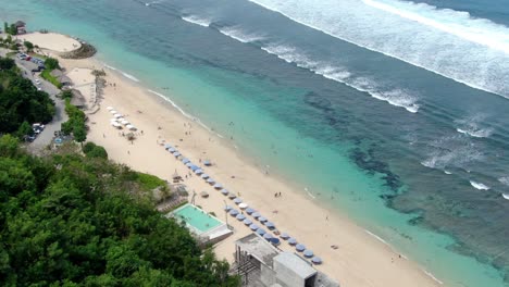 From-the-edge-of-the-hill-in-ULUWATU-BALI,-MELASTI-BEACH-is-visible,-along-with-various-hotels-and-sunshades,-and-a-large-number-of-people-who-are-either-strolling-on-the-beach-or-swimming-in-the-sea