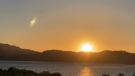 Lens-flare-of-sun-through-huge-river-on-panorama-of-mountain-landscape-at-sunrise-and-bright-disk-of-sun-in-morning-in-summer-against-blue-sky-aerial-view-of-drone-forward