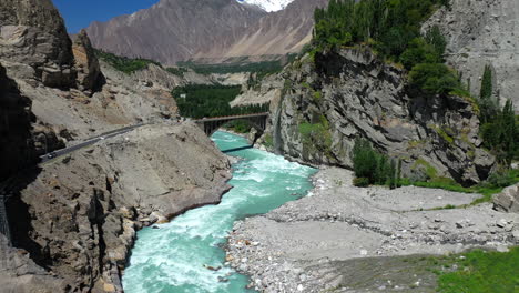 Drone-shot-of-turquoise-blue-water-river-flowing-under-a-bridge-in-Karakoram-mountain-range-along-Karakoram-highway,-moving-through-mountain-pass-towards-bridge