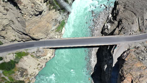 Drone-shot-of-turquoise-blue-water-river-flowing-under-a-bridge-in-Karakoram-mountain-range-along-Karakoram-highway,-rising-aerial-shot