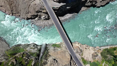 Drone-shot-of-turquoise-blue-water-river-flowing-under-a-bridge-in-Karakoram-mountain-range-along-Karakoram-highway,-downward-angle