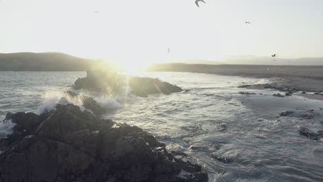 Erstaunliche-Aufnahme-Der-Küste-Eines-Strandes-Während-Des-Sonnenuntergangs-Mit-Fliegenden-Möwen,-Während-Die-Wellen-Gegen-Die-Felsen-Prallen-Und-Die-Sonne-Das-Meer-Erleuchtet