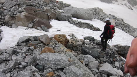 A-crystal-seeker-mountaineer-walks-on-rocks-next-to-a-snow-plain,-he-has-all-his-equipment,-helmet-and-pickaxe,-swiss-alps