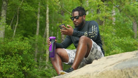 A-man-sits-down-and-reads-his-phone-on-a-giant-rock-in-the-middle-of-the-woods
