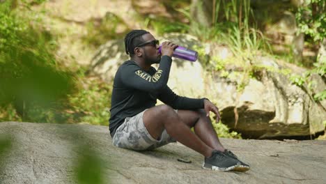 A-man-takes-a-sip-of-water-from-a-bottle-on-a-giant-rock-in-the-middle-of-the-woods
